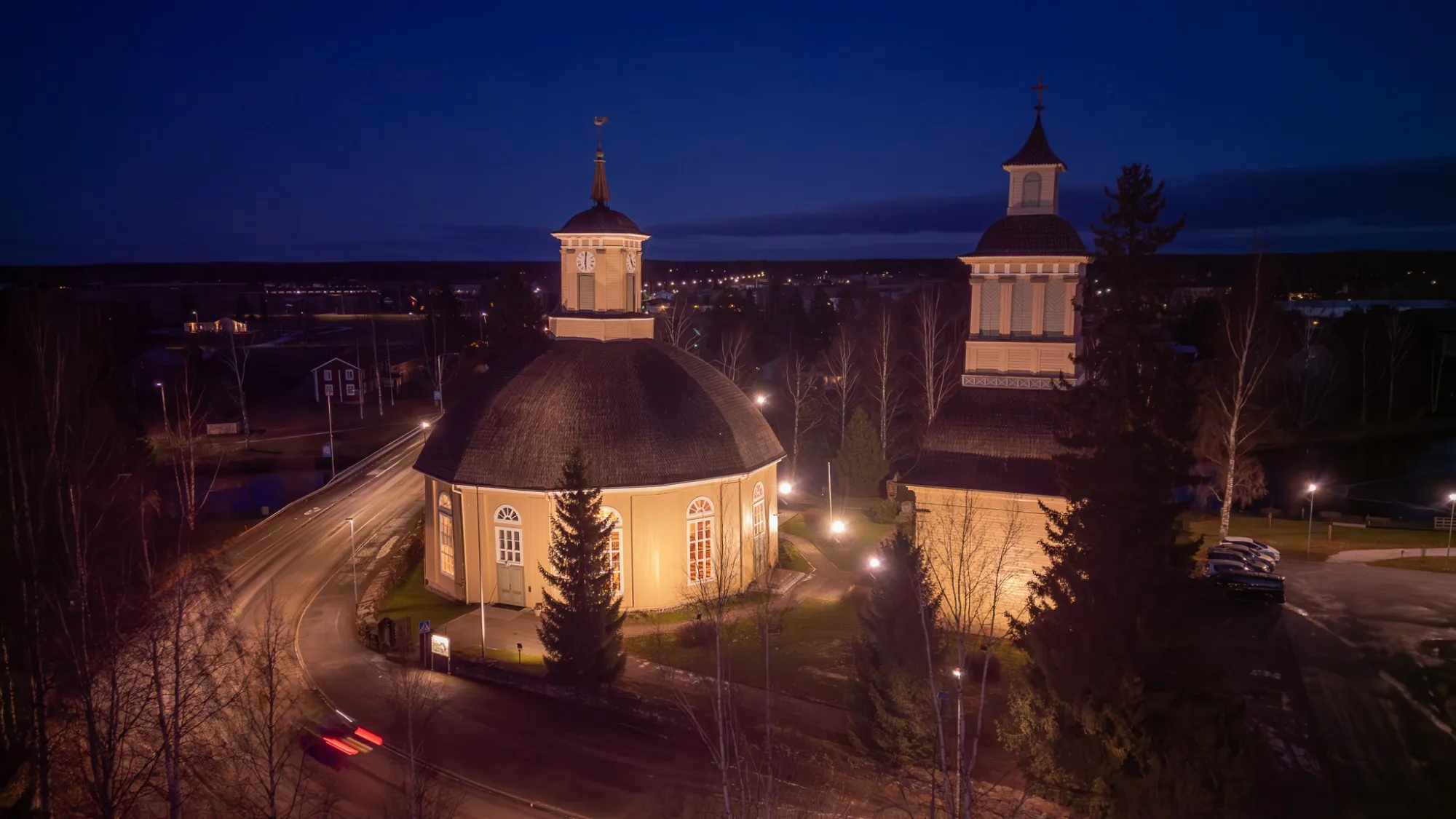 Ilmakuva Vimpelin kirkosta ja kellotapulista iltahämärässä, auton valot piirtävät juovan tielle